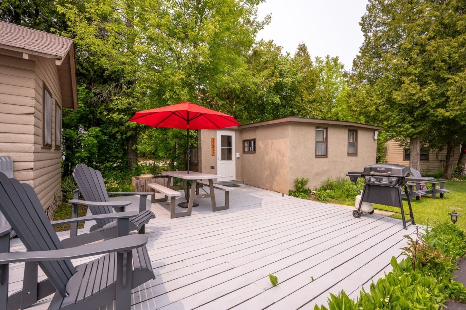 Wooden deck with Adirondack chairs, picnic table, red umbrella, and a grill. Small buildings and trees.