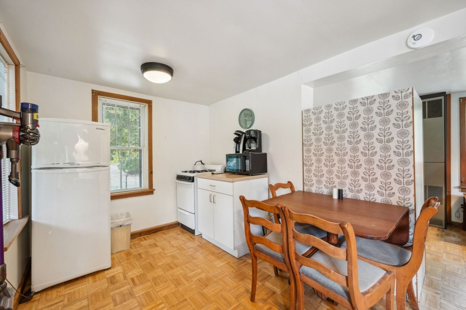 Small kitchen with a white refrigerator, stove, and cabinets. A wooden table and chairs are in the center.