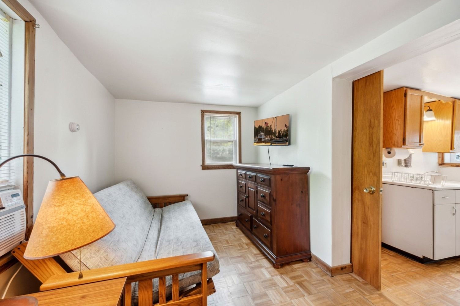 Small room with a futon, wooden dresser, and a doorway to a kitchen area with cabinets.
