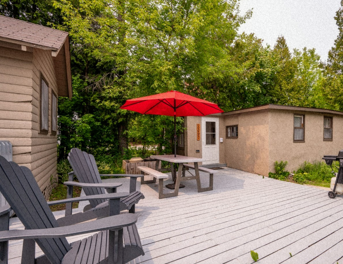 Deck with Adirondack chairs, picnic table under a red umbrella, two cabins, and trees.