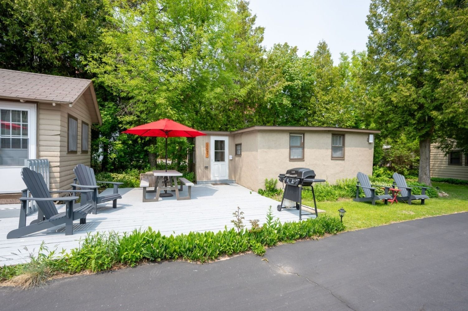Exterior of a cabin with patio, table, chairs, grill, and red umbrella. Green trees in the background.