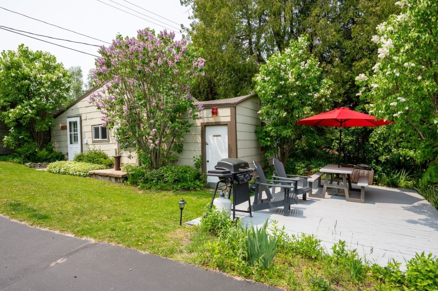 Cottage with grilling area: light yellow building, red umbrella, grill, patio with seating, lilac bush, green lawn.
