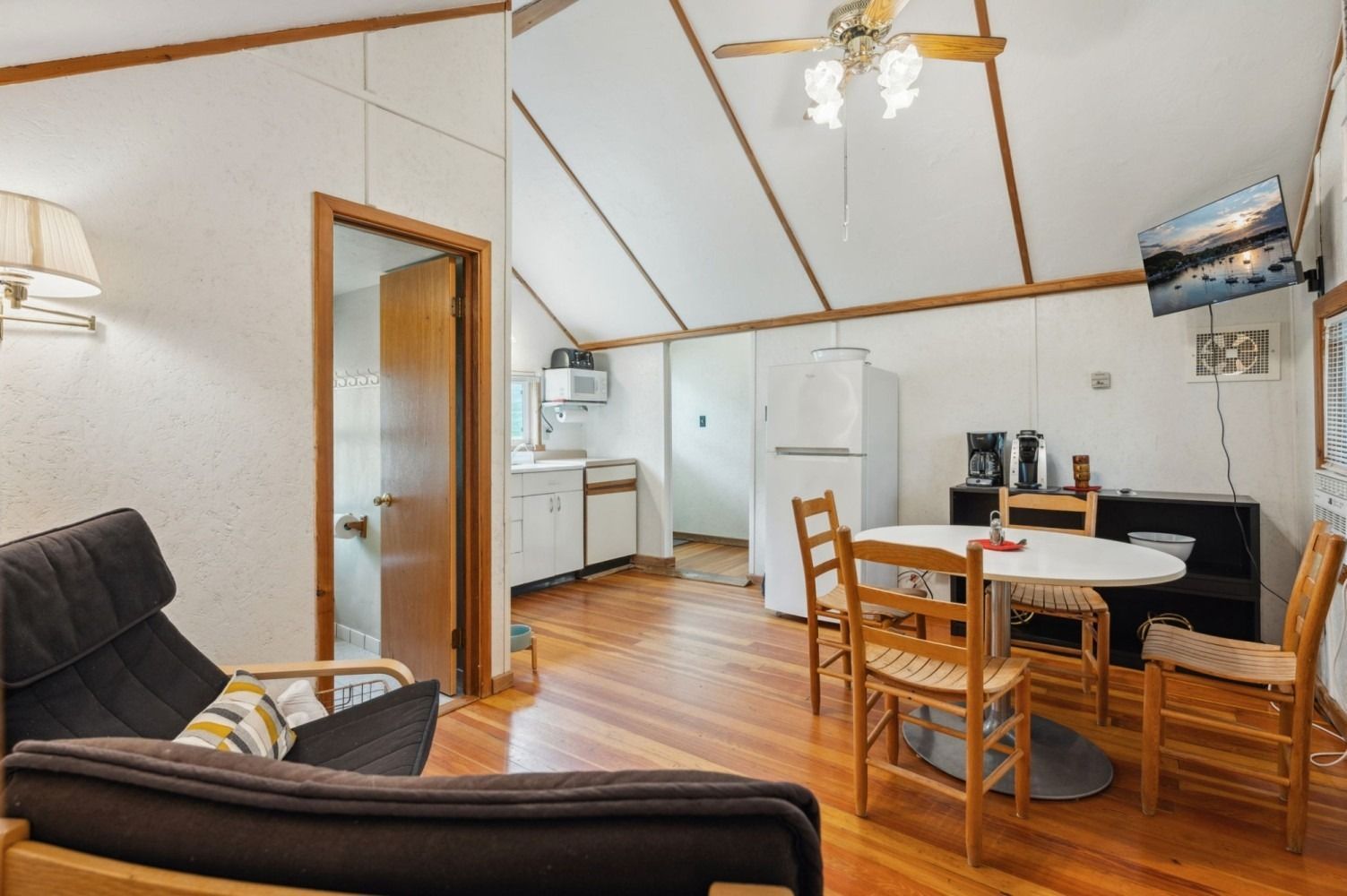 Cozy cabin interior with kitchen, dining area, and seating. Wooden floors, white walls, and a ceiling fan.