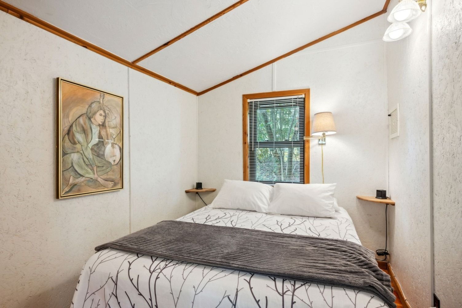 Small bedroom with bed, artwork, window, and wall-mounted lamp.