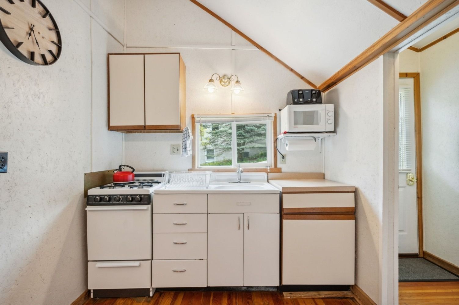Small white kitchen with stove, sink, cabinets, microwave, and red kettle; wood floors, window, and clock on wall.