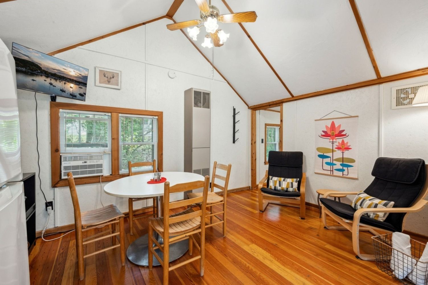 Small cabin interior with table, chairs, armchairs, and a TV; hardwood floors.