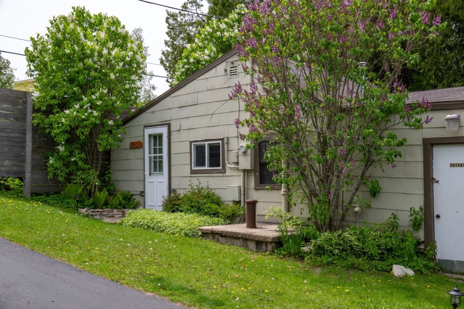 Small beige building with a white door, a small window, and blooming trees. Green grass slopes up to the structure.