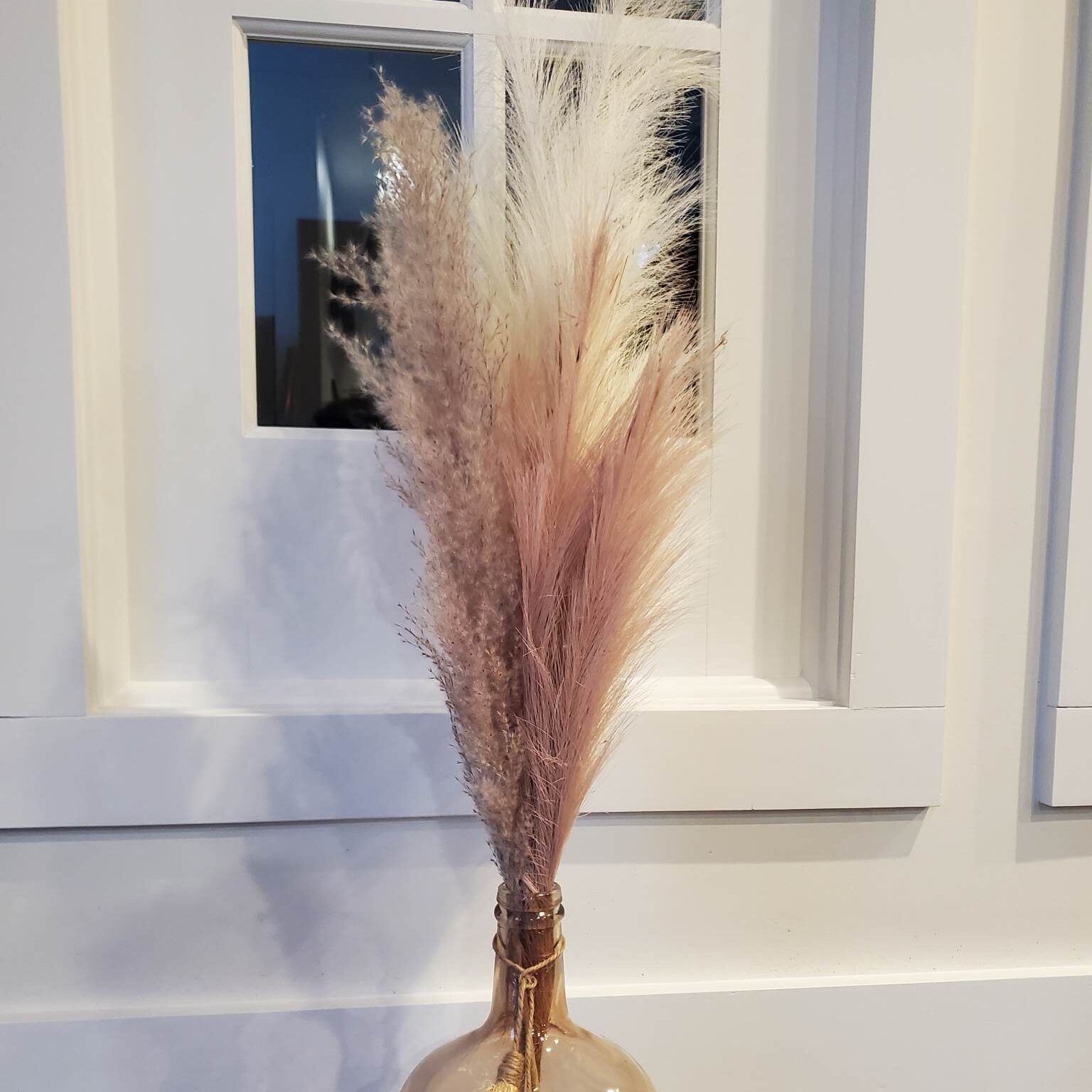 Pampas grass arrangement in a blush-colored glass vase, set in front of a white framed window.