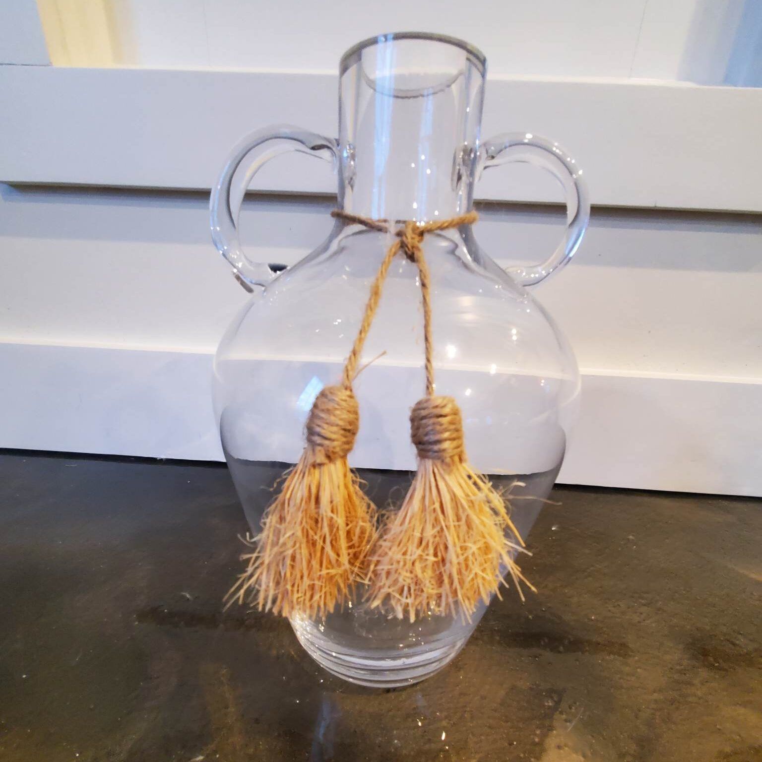 Clear glass vase with two handles, tied with a jute rope and tassels.