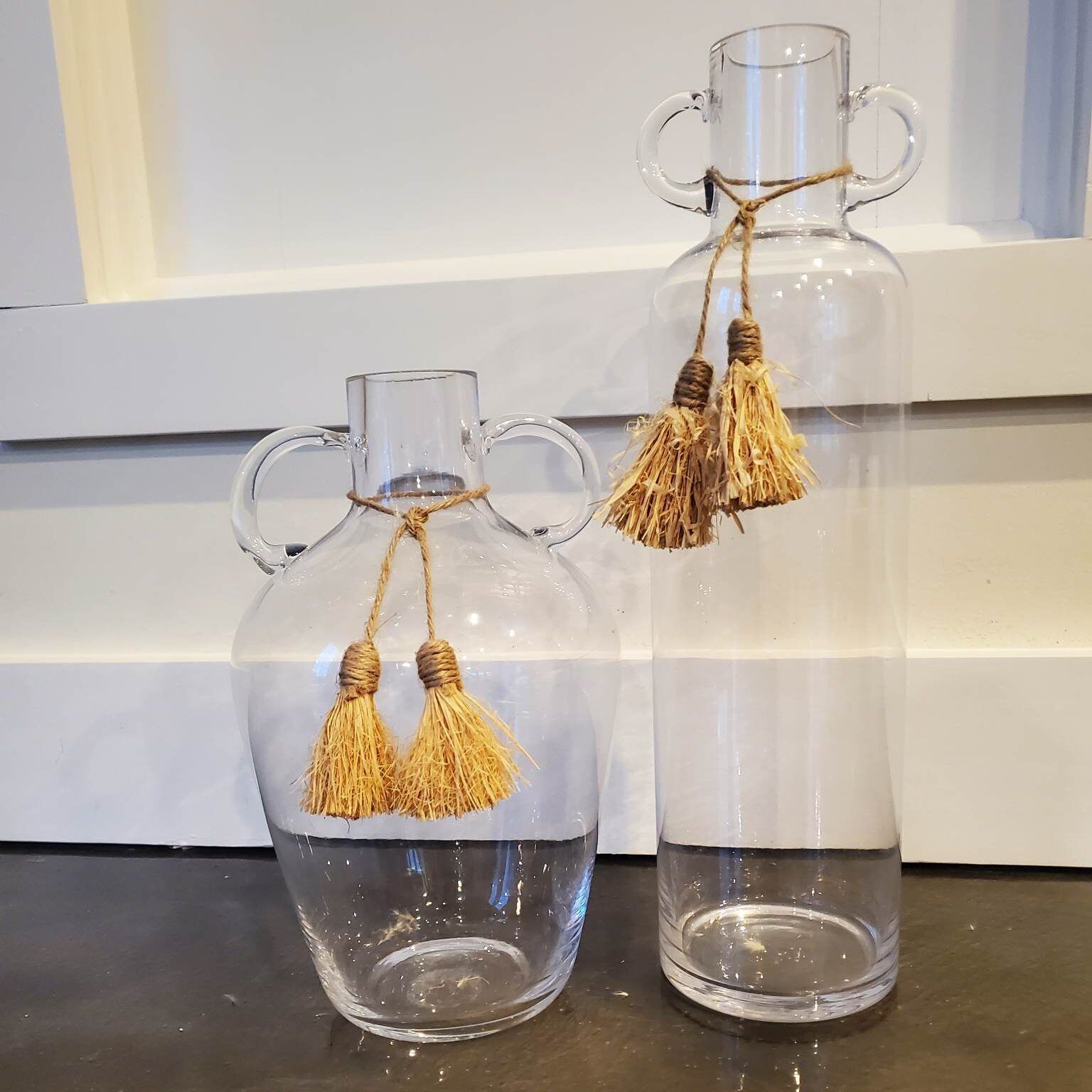 Two clear glass vases with handles and decorative tassels, on a dark surface against a white wall.