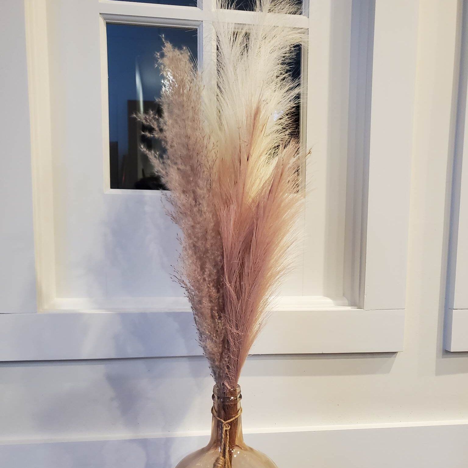 Pink and white pampas grass in a peach-colored vase, set against a white window frame.