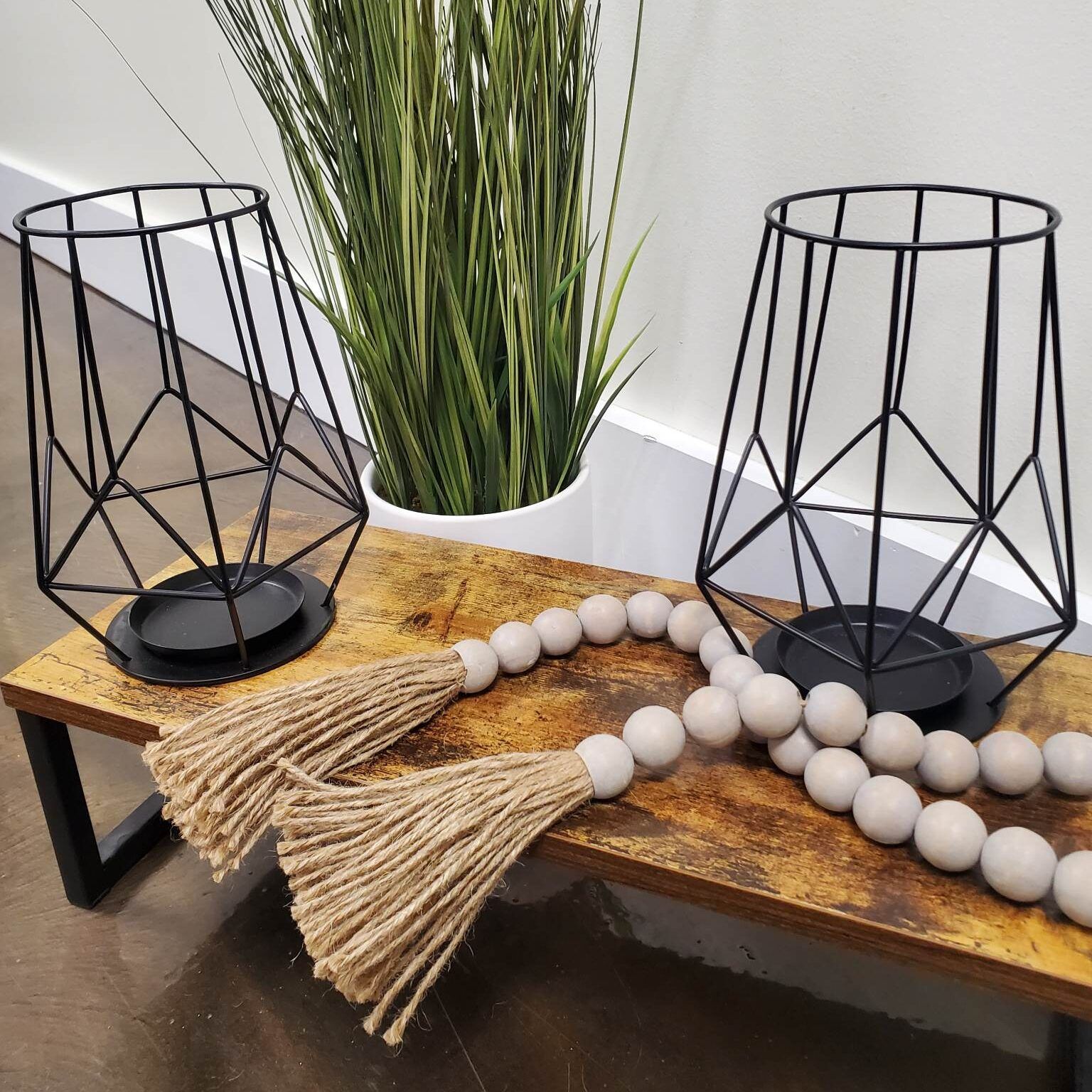 Two black geometric candle holders, wooden beads, and faux greenery on a rustic bench.