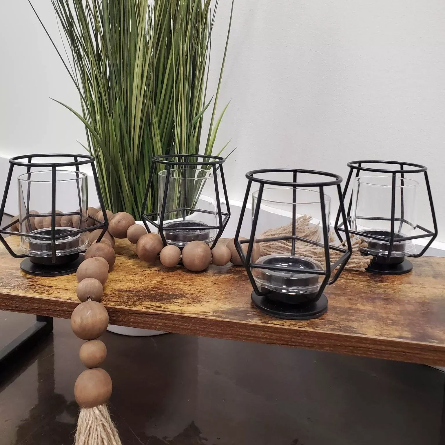 Four black metal candle holders on a wooden shelf, with greenery and wooden beads.
