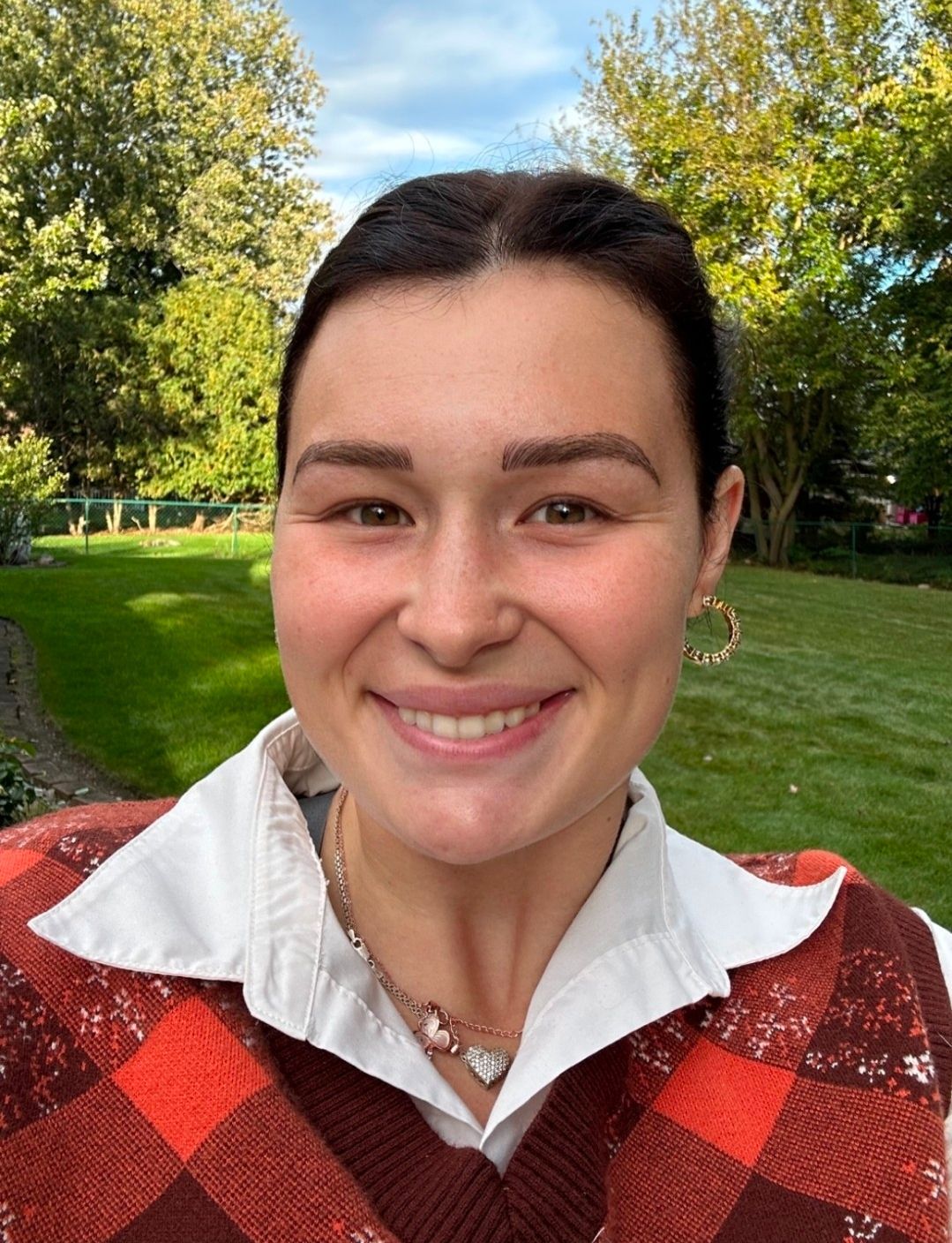 Woman smiles at the camera outdoors, wearing a white collared shirt under a red and brown sweater.