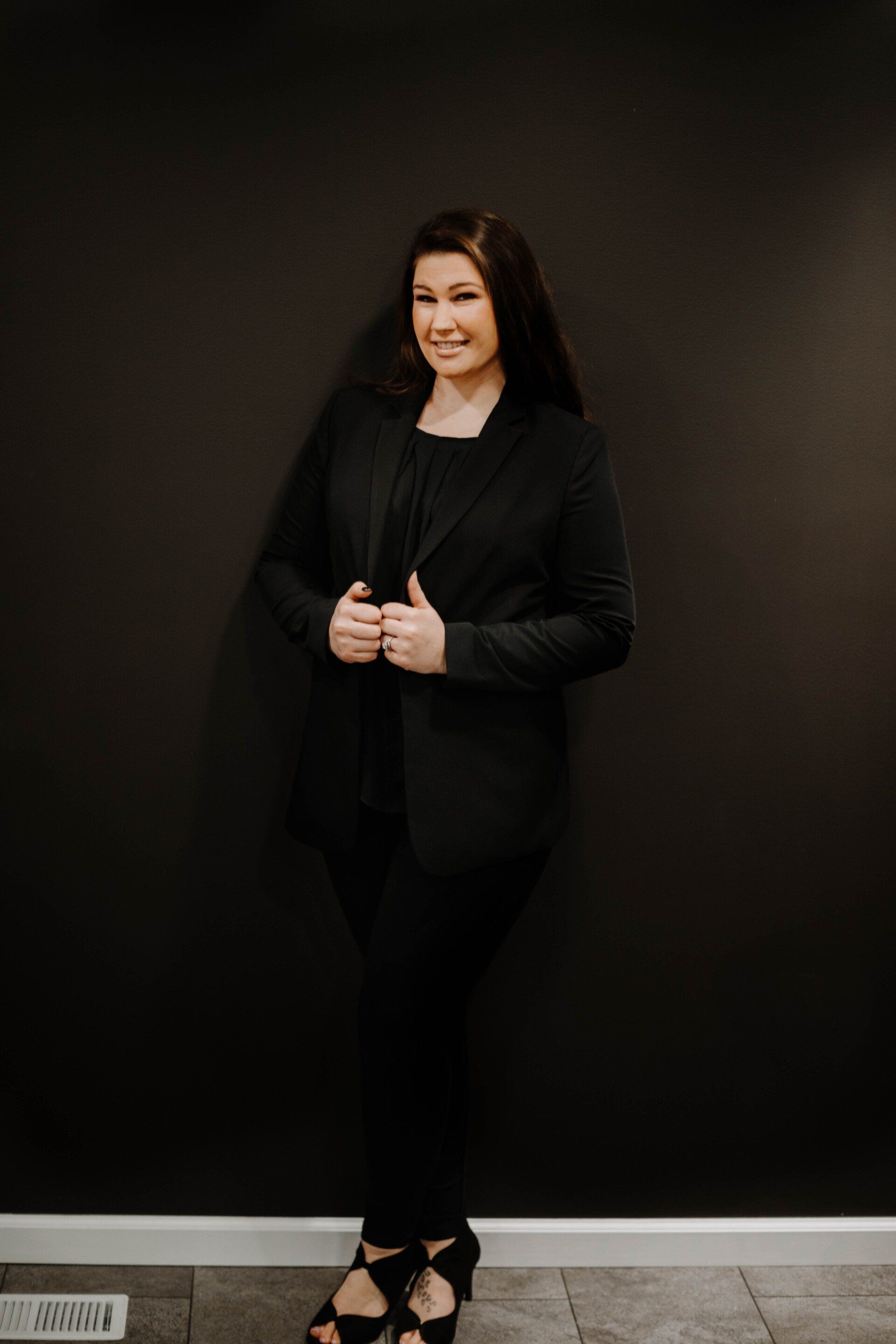 Woman in black blazer and pants, smiling, leaning against a dark wall.