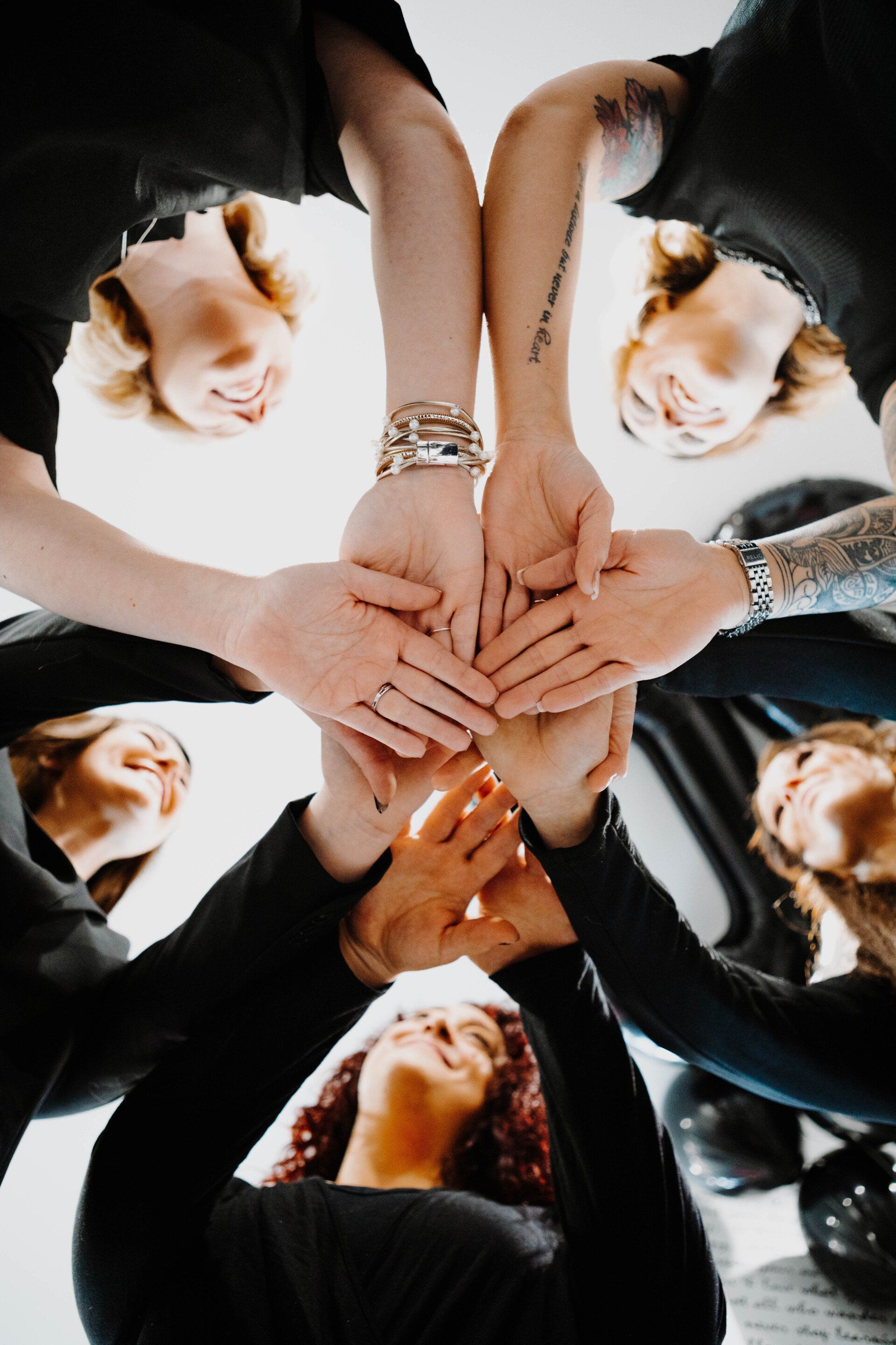Hands of multiple people touching in a circle; faces looking up from below, all in black.