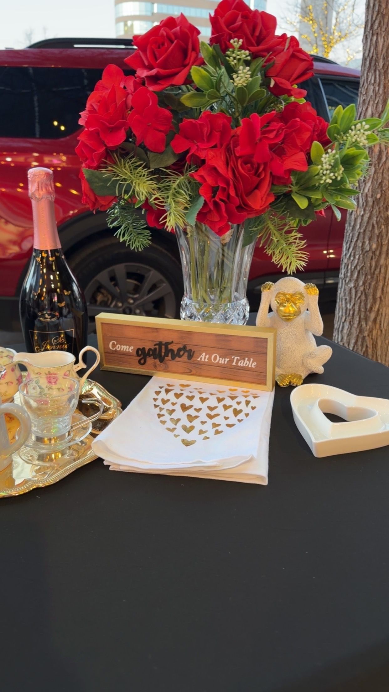 A bouquet of red roses, a bottle of pink sparkling wine, and decor with hearts on a black table with a red car backdrop.