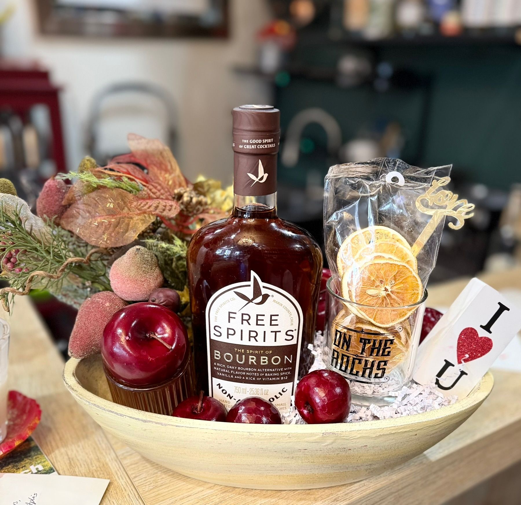 A gift basket featuring a bottle of Free Spirits bourbon, dried orange slices, a tumbler, and apples in a beige bowl.
