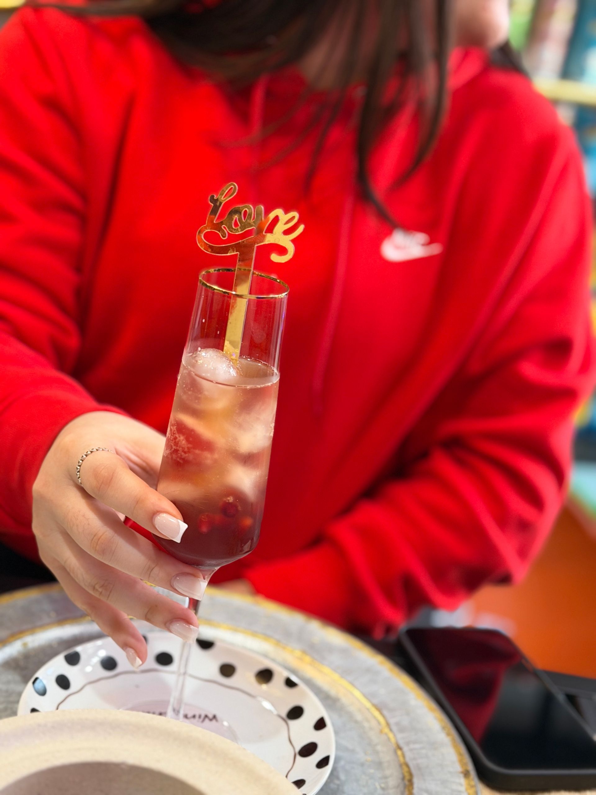 A person in a red hoodie holds a cocktail in a champagne flute featuring a gold 