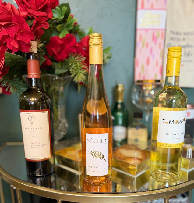 Three wine bottles stand on a gold-rimmed glass table in front of a green wall and a vase of red flowers.