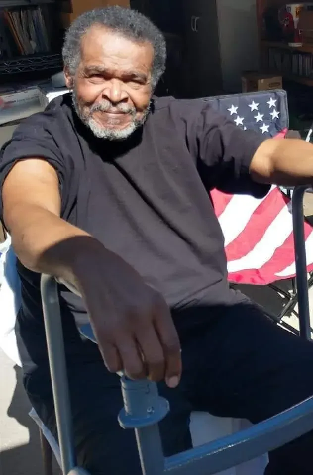 Man in black shirt sits in a folding chair, smiling. 