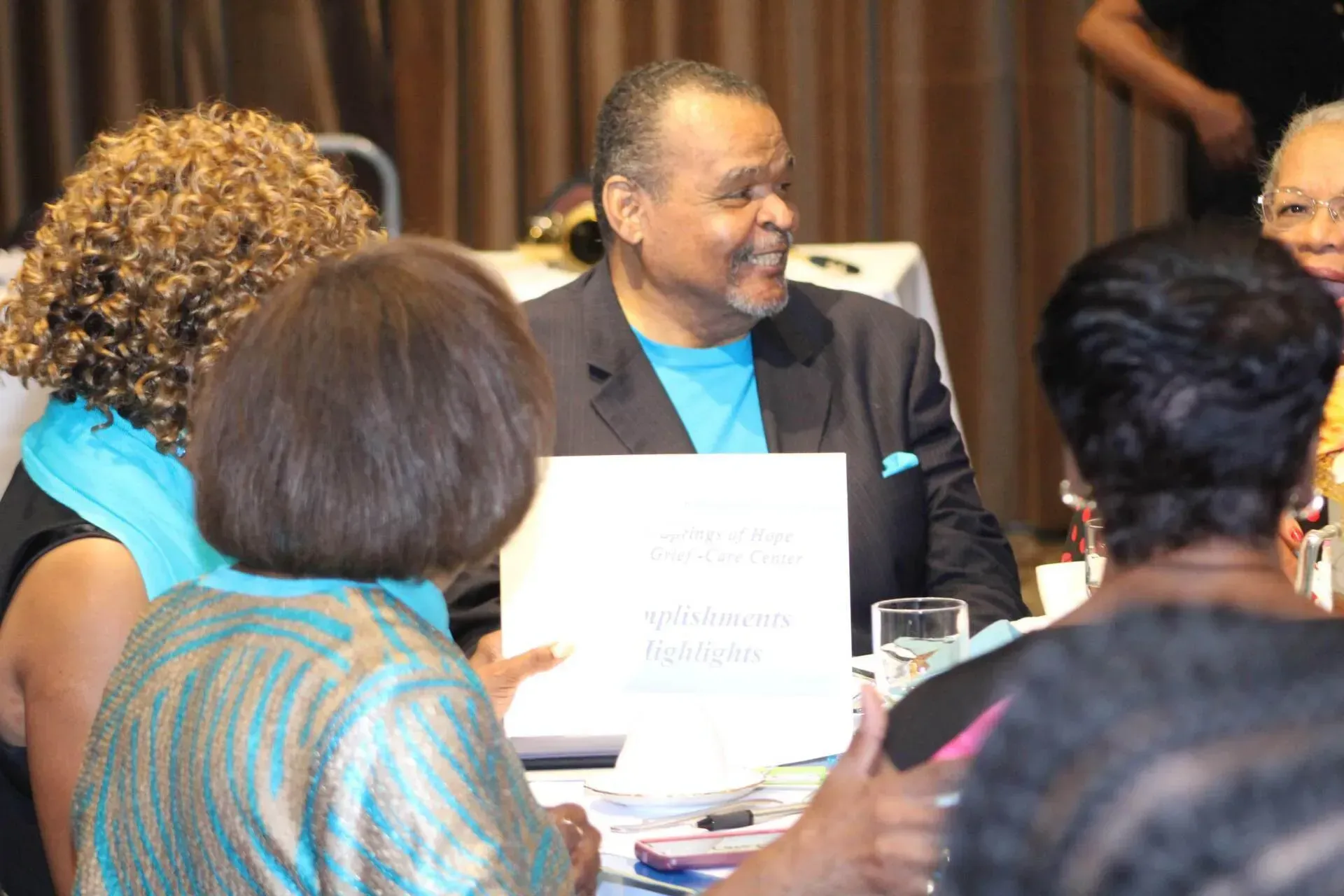 Man in suit smiles at a table with other people; holds a sign. Indoor, daytime.