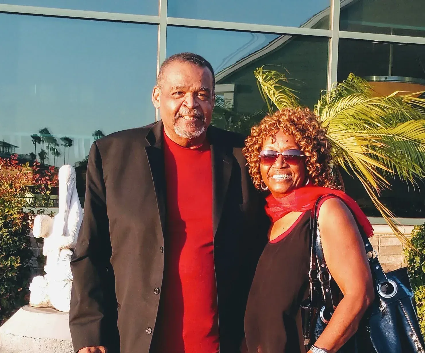 A couple poses outdoors near a building with windows; man in black jacket and red shirt; woman in black top and red scarf.