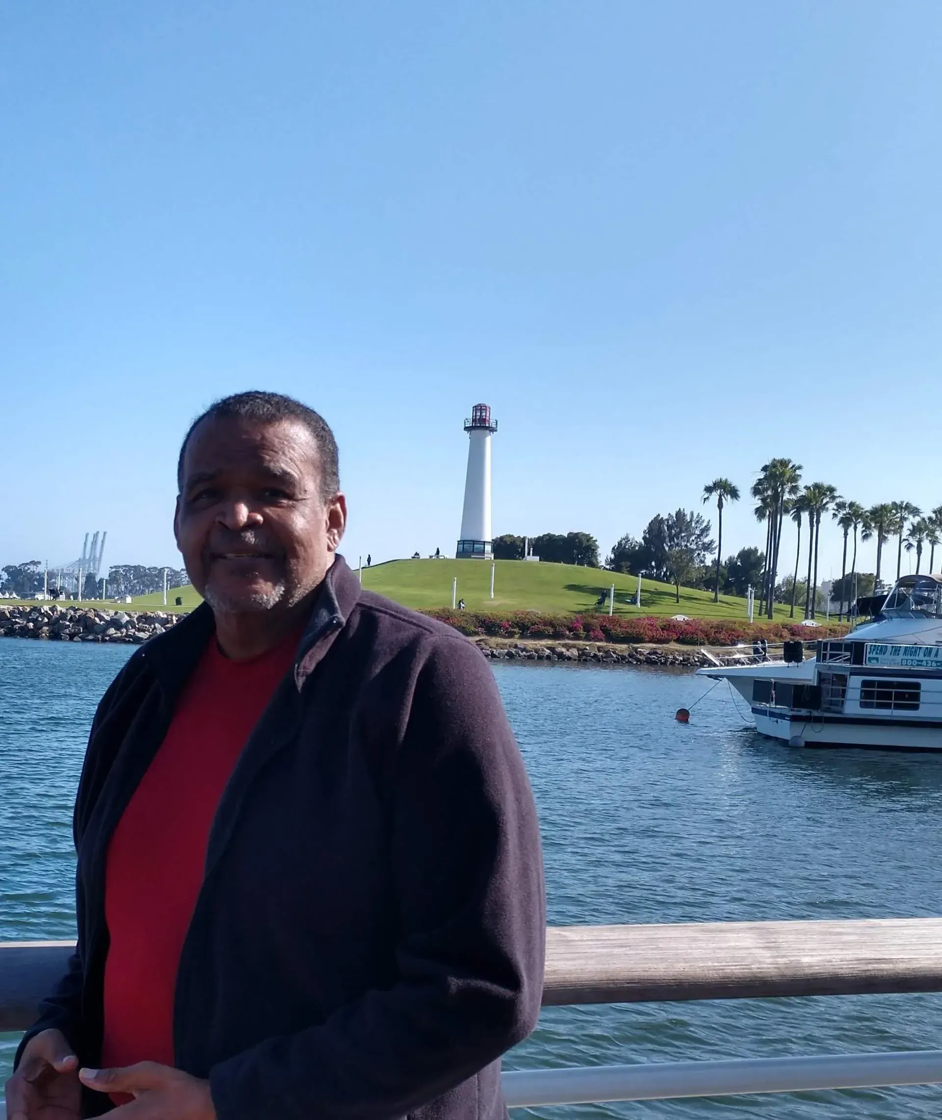 Man stands in front of a waterfront with a lighthouse in the distance; sunny, blue sky.