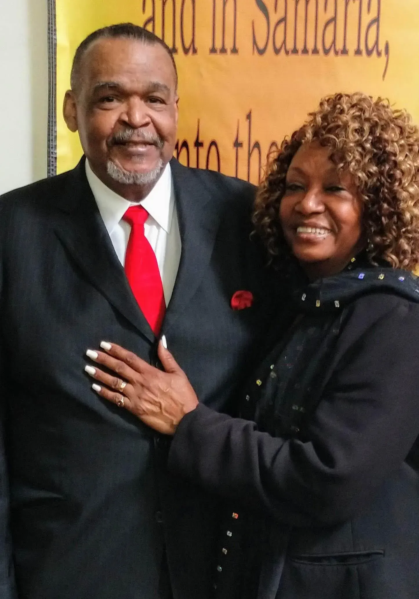 A man in a suit and a woman in a coat pose together, smiling, with one hand on his chest.