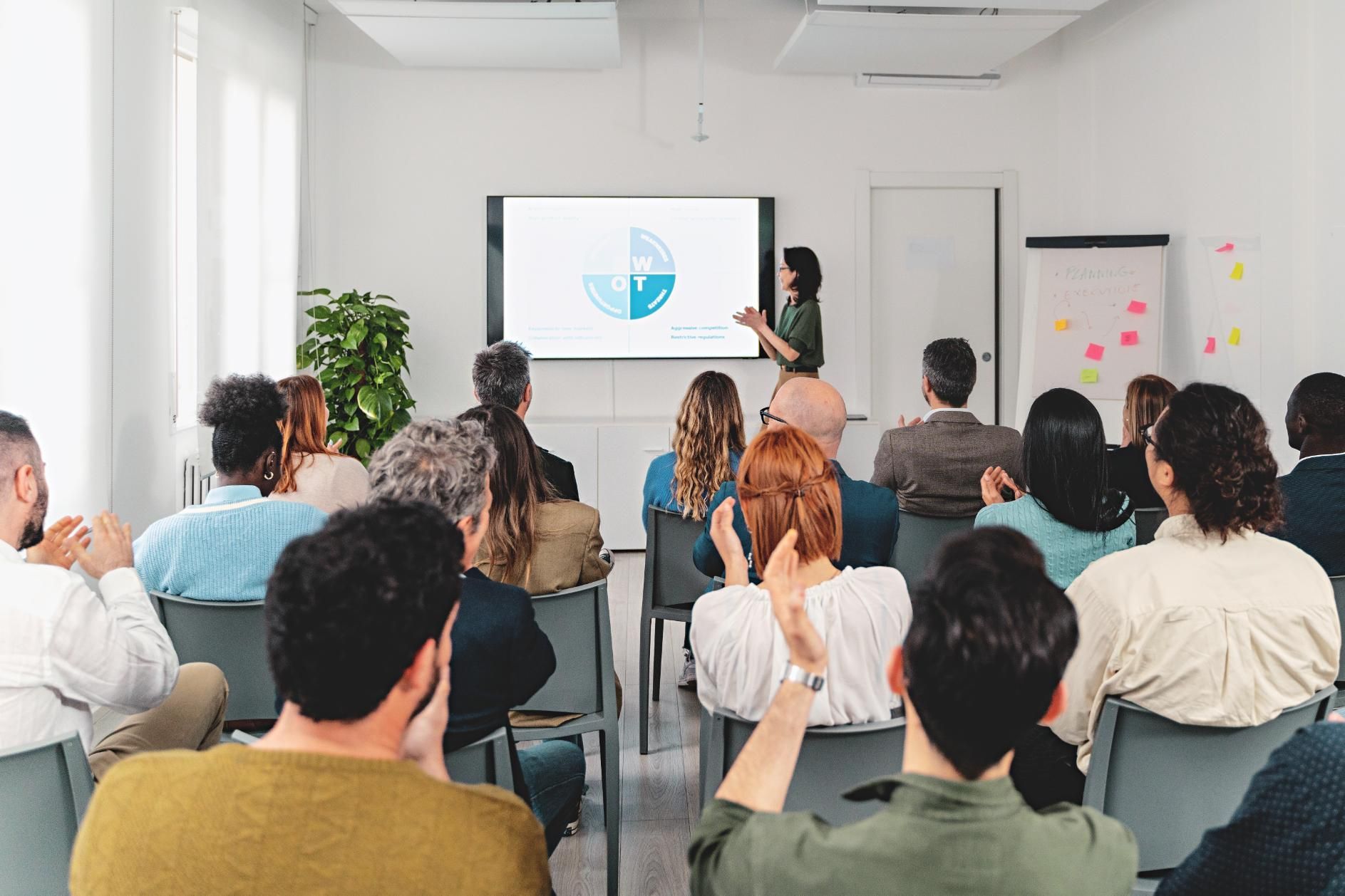 Woman presenting to an audience, displaying a pie chart on a screen in a conference room. 