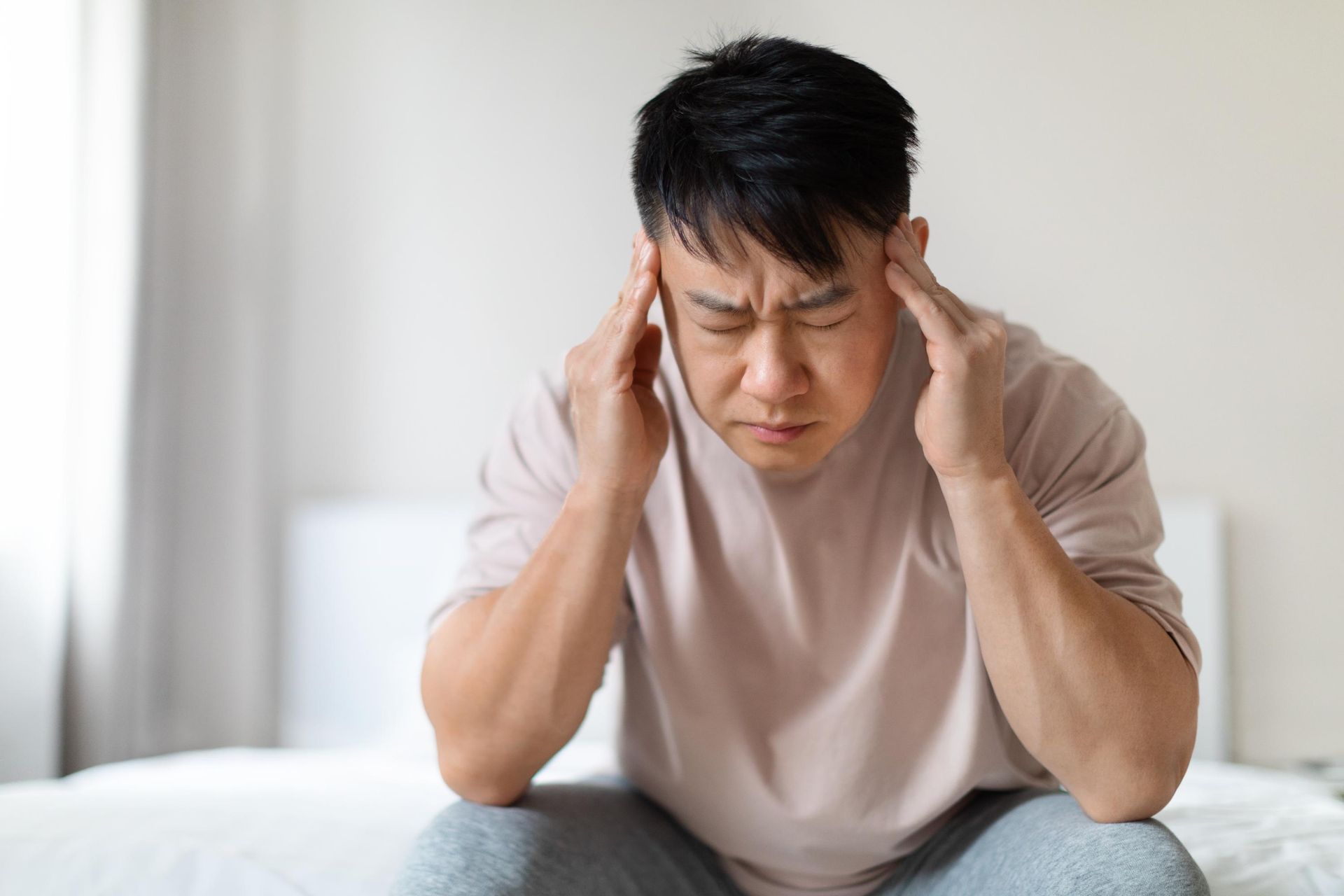 Man sitting, clutching head in pain, eyes closed. Indoors, neutral background.