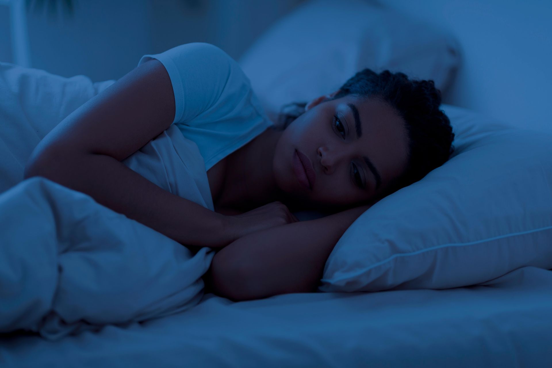Woman in bed, lying down, looking thoughtful, illuminated by blue light.