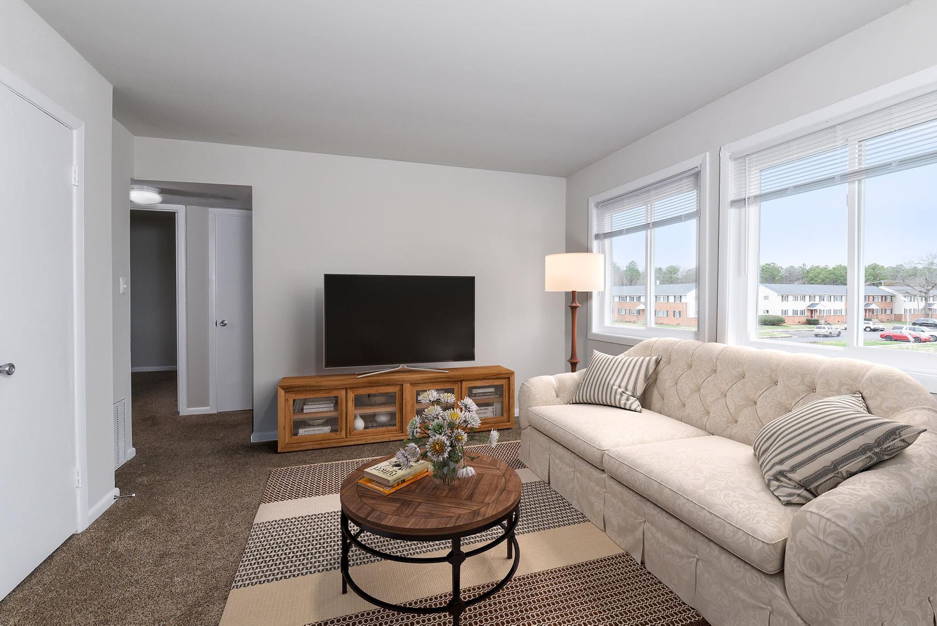 Living room with beige tufted sofa, round coffee table, TV on a wooden console, and large windows.