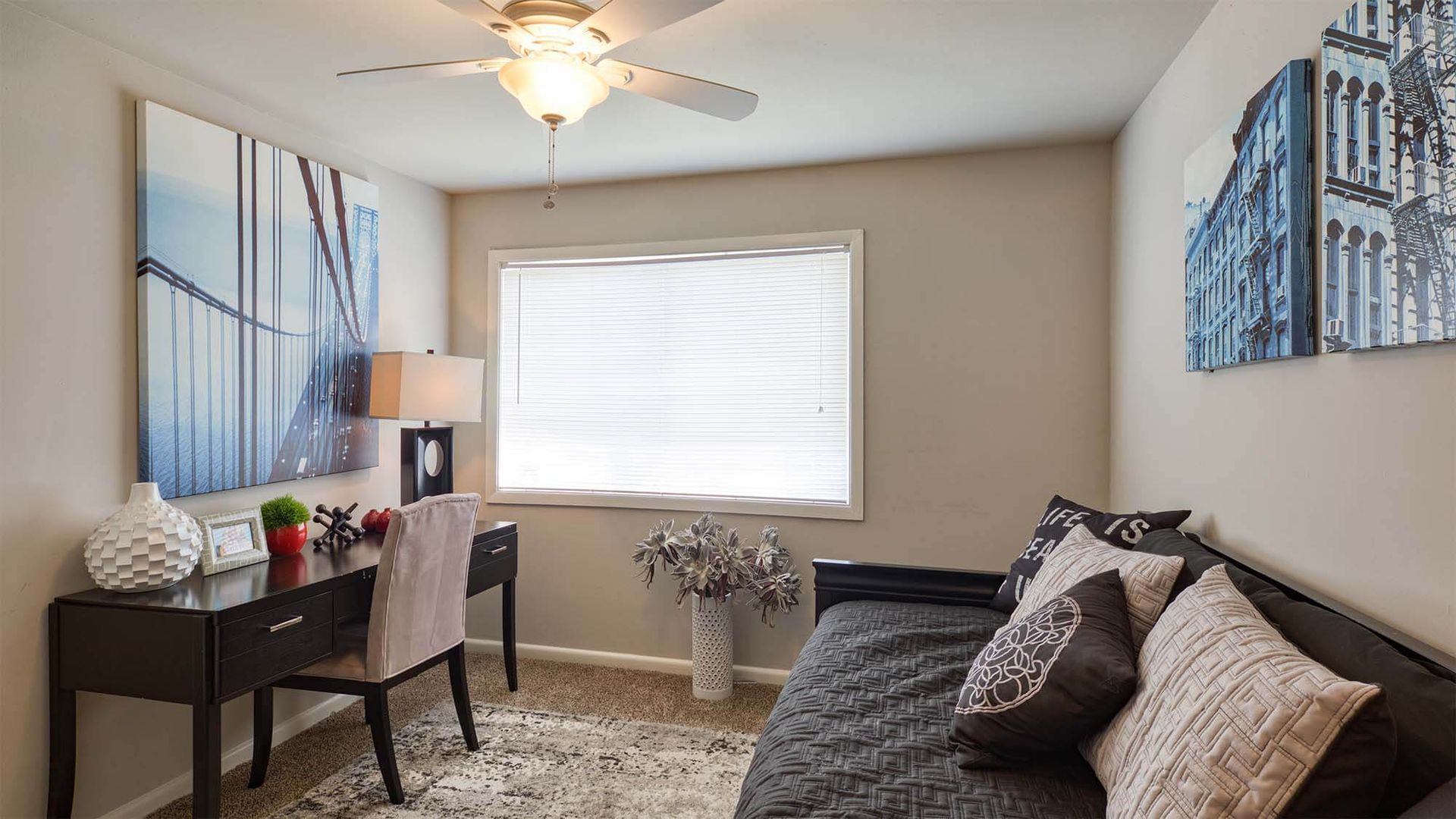 Bedroom with desk and daybed, window with blinds, and two cityscape paintings.