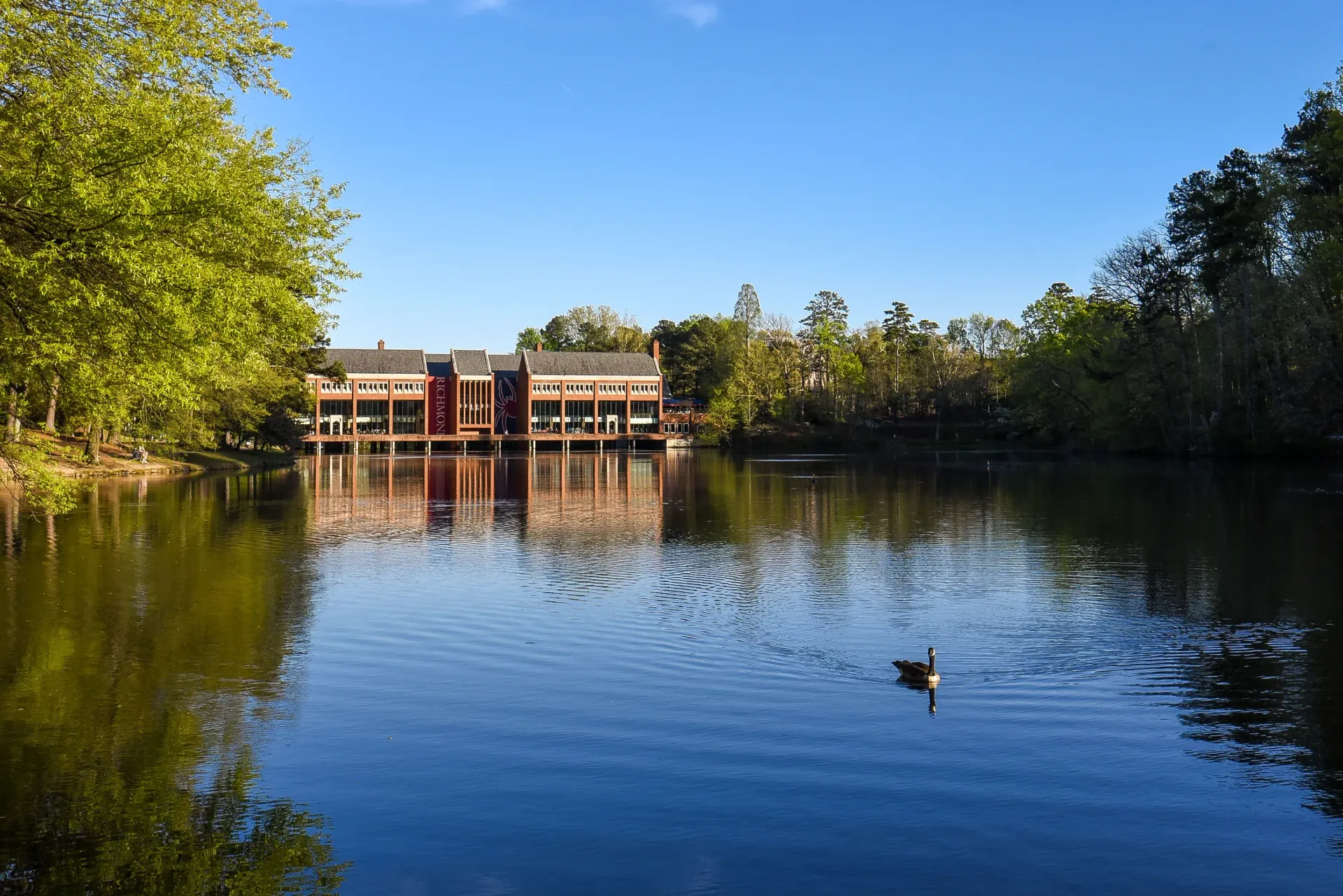 Lakeside brick community building with banners along a calm lake and trees.