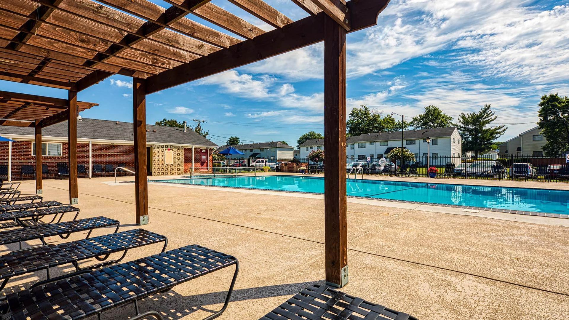 Outdoor community pool under a wooden pergola with rows of lounge chairs.