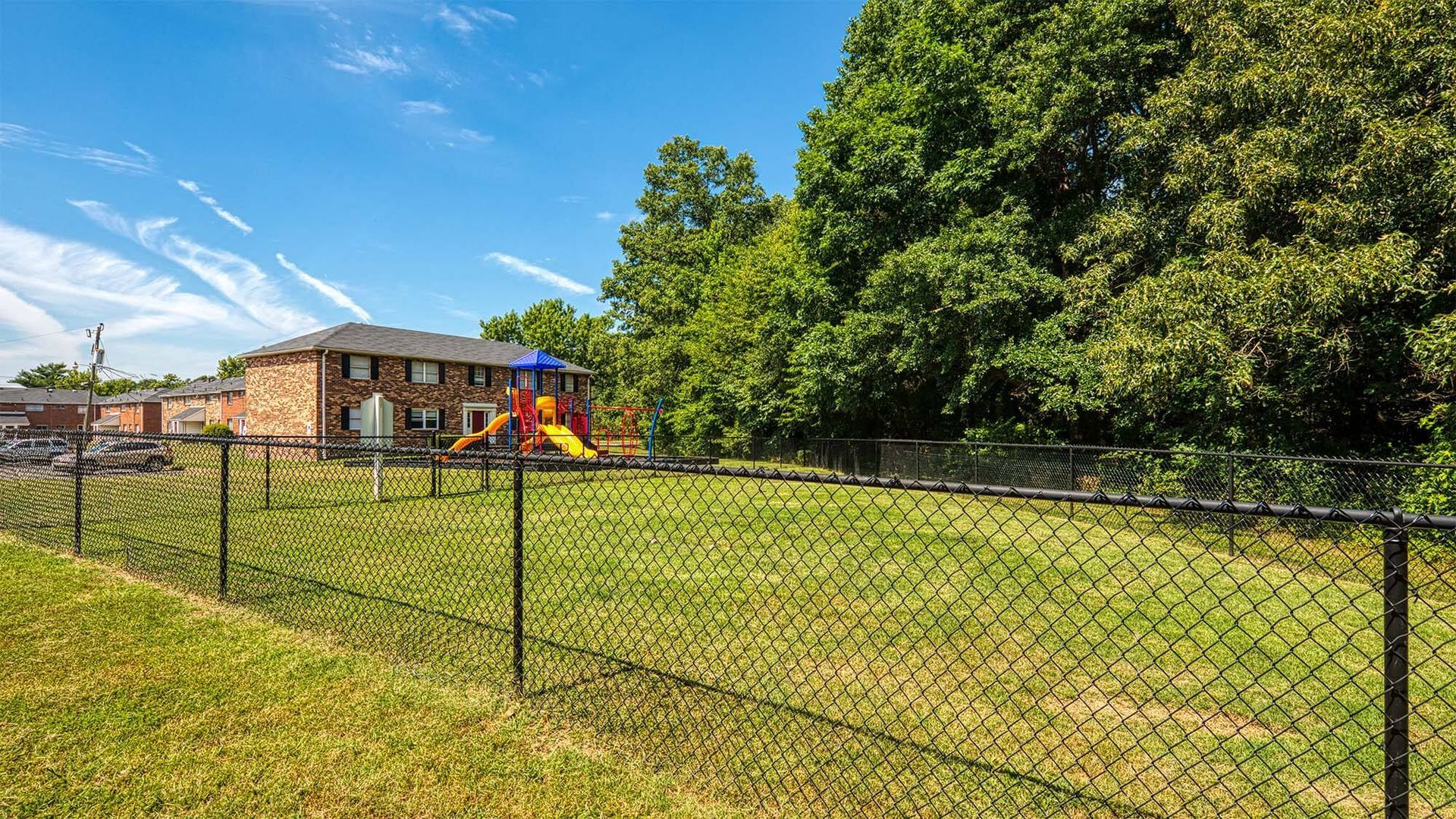 Fenced grassy playground with a colorful play structure, brick apartment buildings in the background.