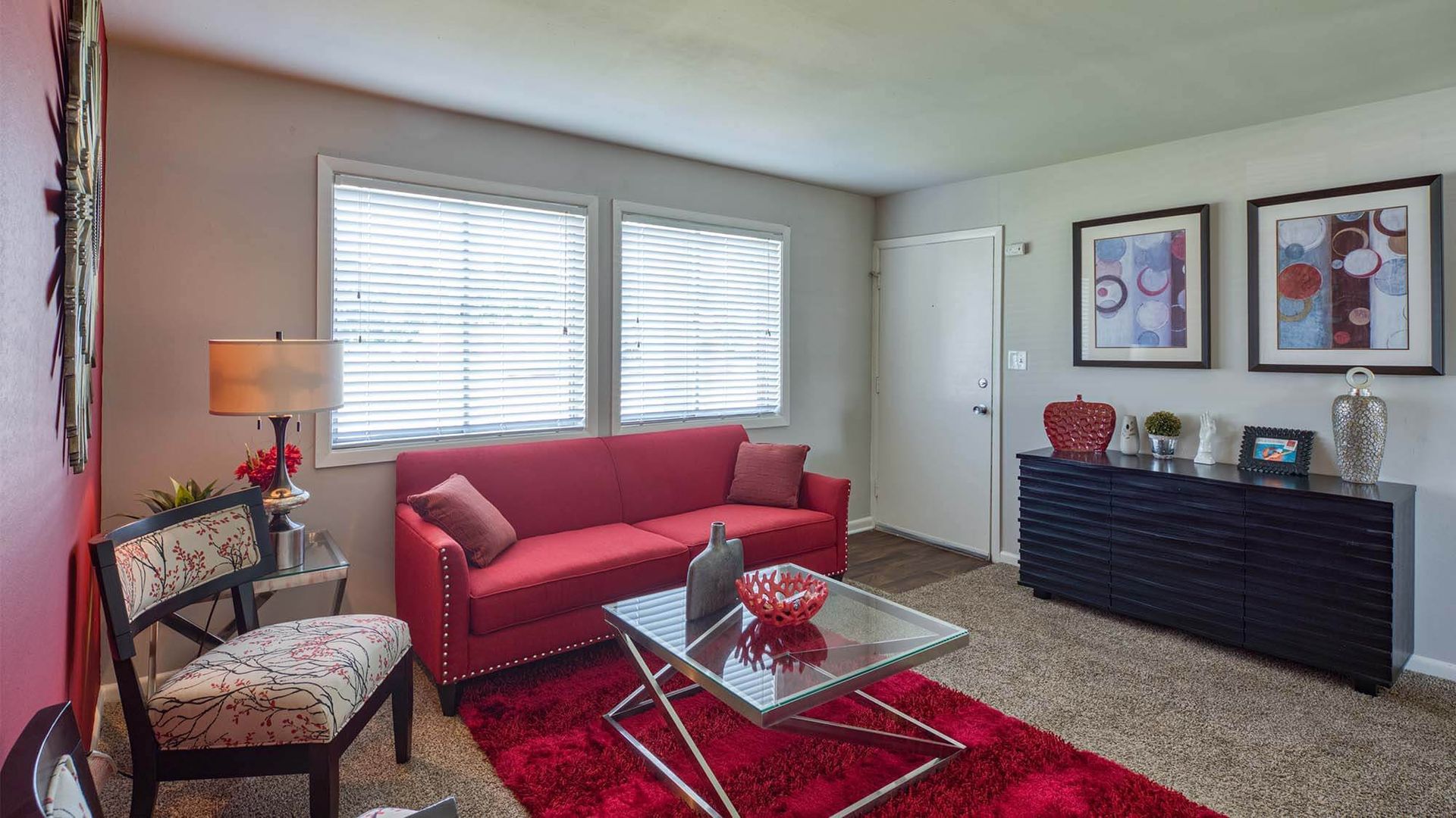 Living room with red sofa, glass coffee table, two bright windows with blinds, and a dark sideboard.