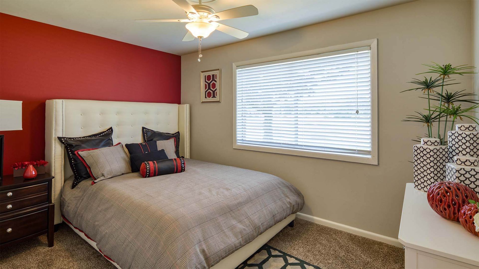Bedroom with white tufted headboard bed, red accent wall, window with blinds, and ceiling fan.