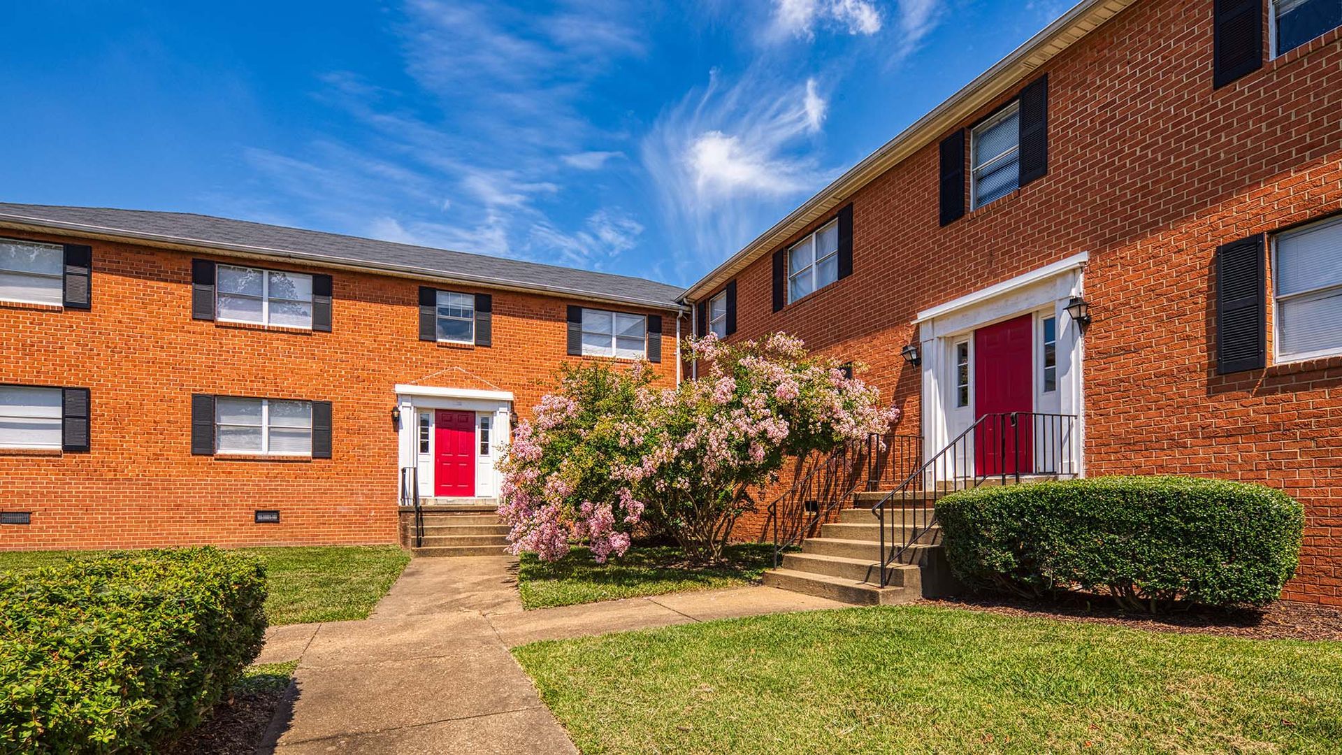 Brick apartment building exterior with red doors, stairs, and flowering shrubbery along a walkway.