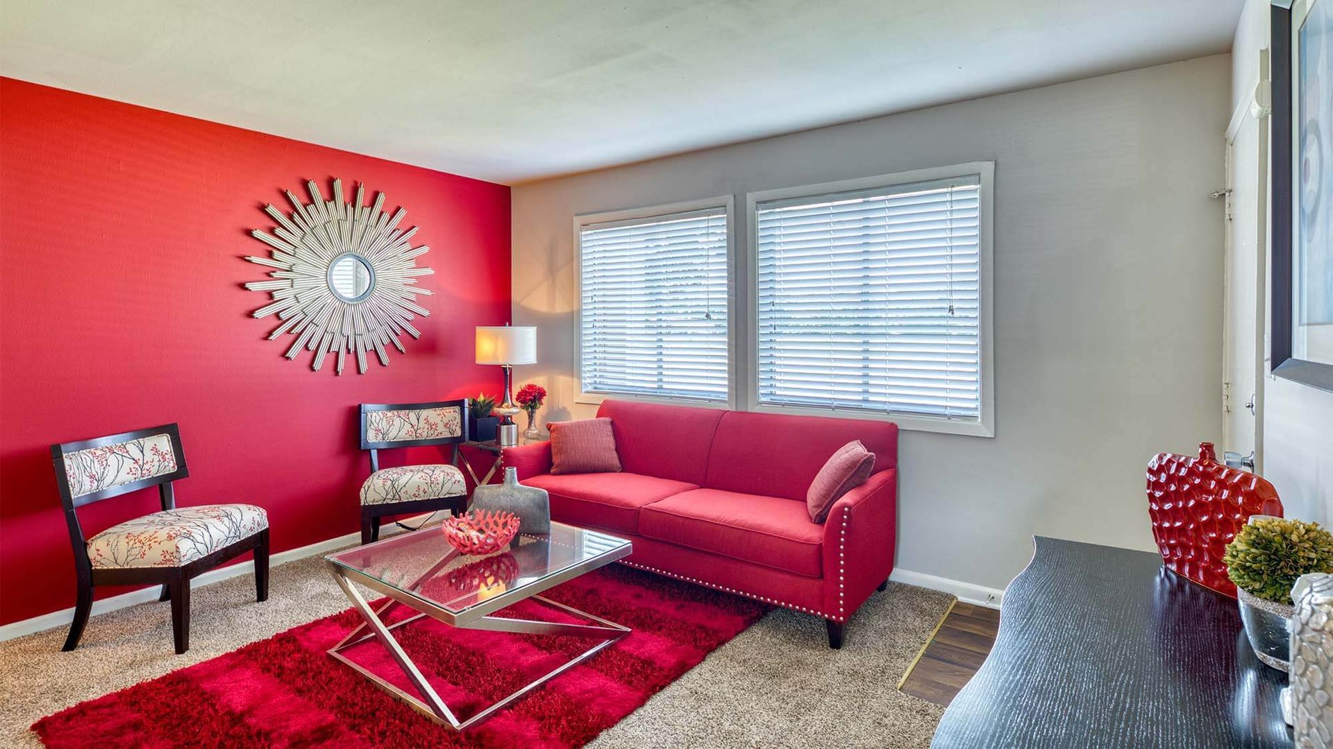 Living room with red accent wall, red sofa, two patterned chairs, and a glass coffee table.