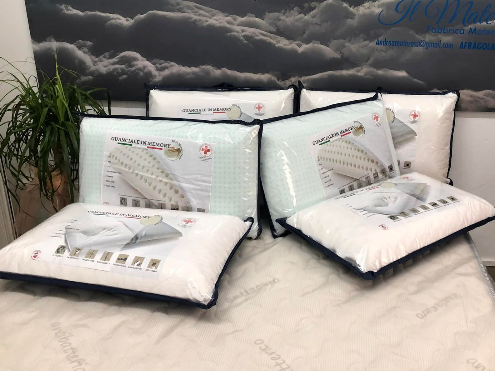Cinque cuscini bianchi da letto, confezionati in plastica trasparente, disposti su un materasso davanti a uno sfondo con un motivo a nuvole.