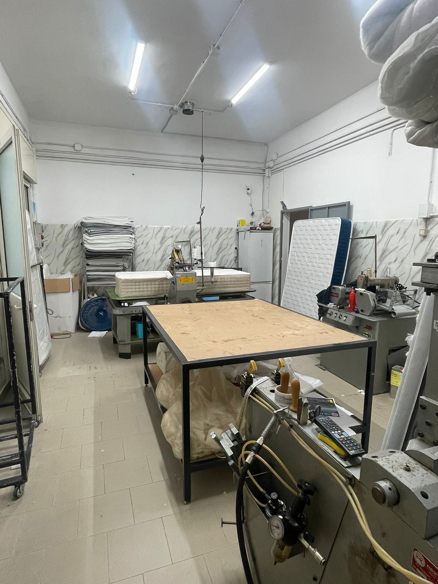 Un laboratorio di produzione di materassi molto luminoso, con macchinari pesanti, grandi tavoli da lavoro in legno e materiali tessili.