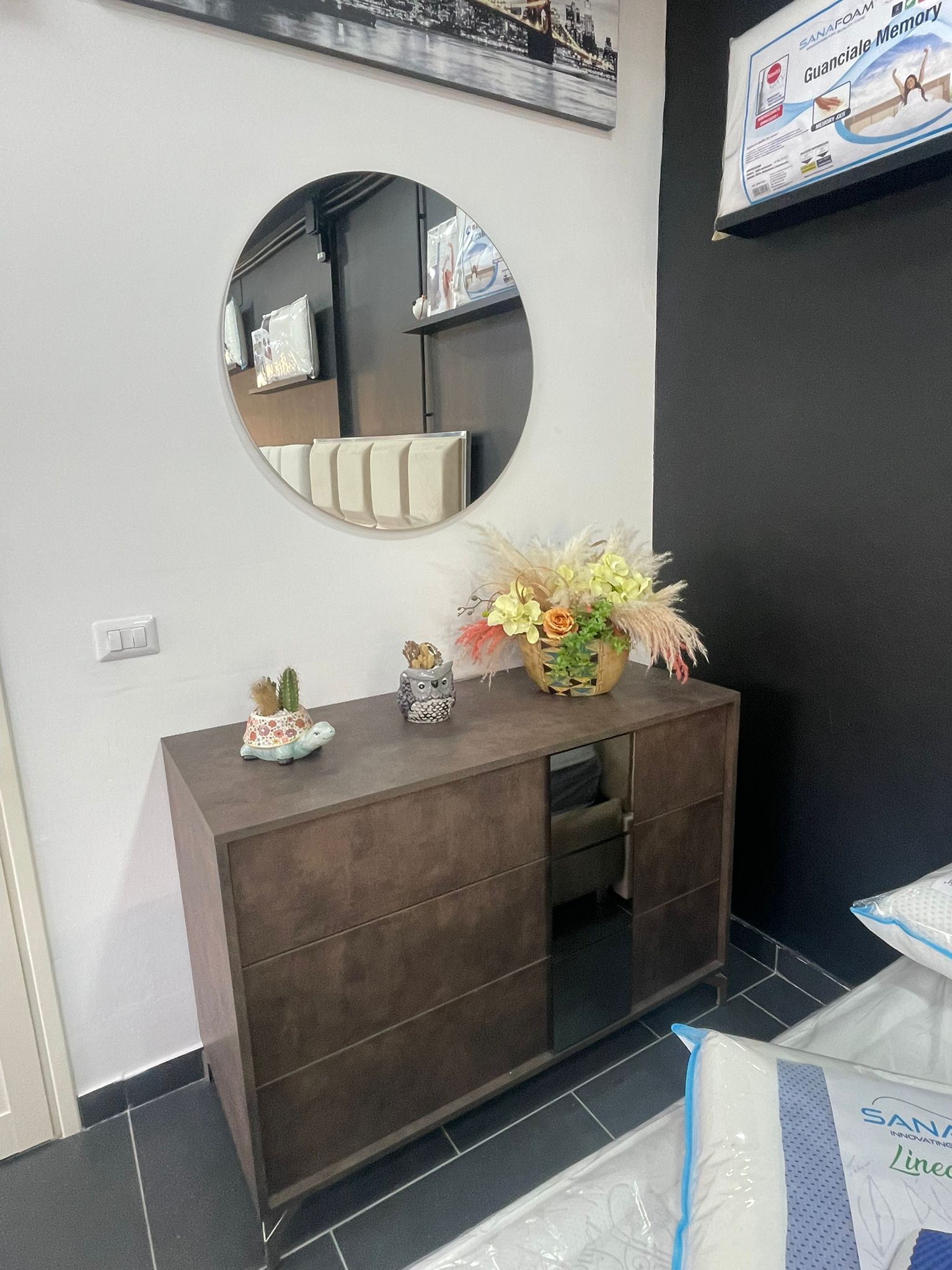 Una cassettiera marrone con uno specchio rotondo sopra e un piccolo mazzo di fiori sopra, situata in una stanza con pavimenti piastrellati scuri.