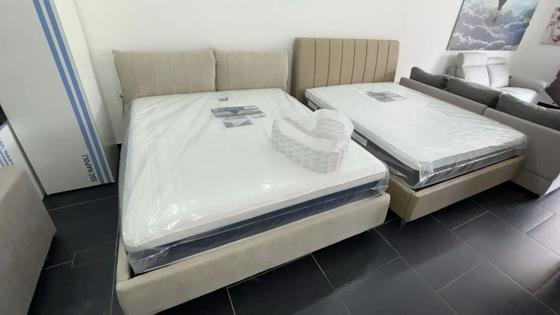 Due letti imbottiti beige con materassi imbottiti sono posizionati uno accanto all'altro in uno showroom di mobili con pavimenti piastrellati scuri.