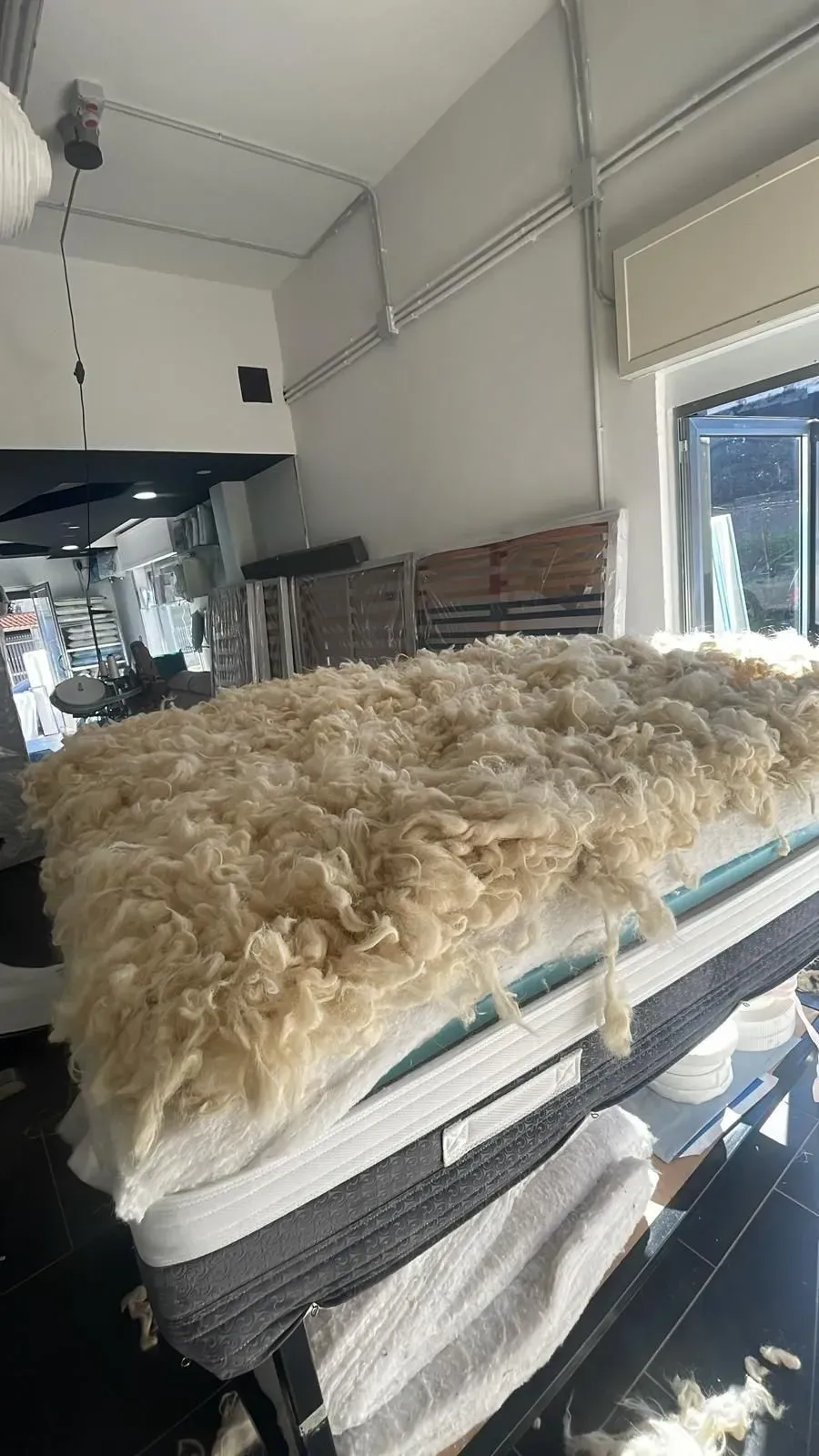 Uno strato di soffice e grezza lana di pecora è adagiato sopra un materasso all'interno di una stanza, mentre nelle vicinanze sono visibili altri componenti del materasso.