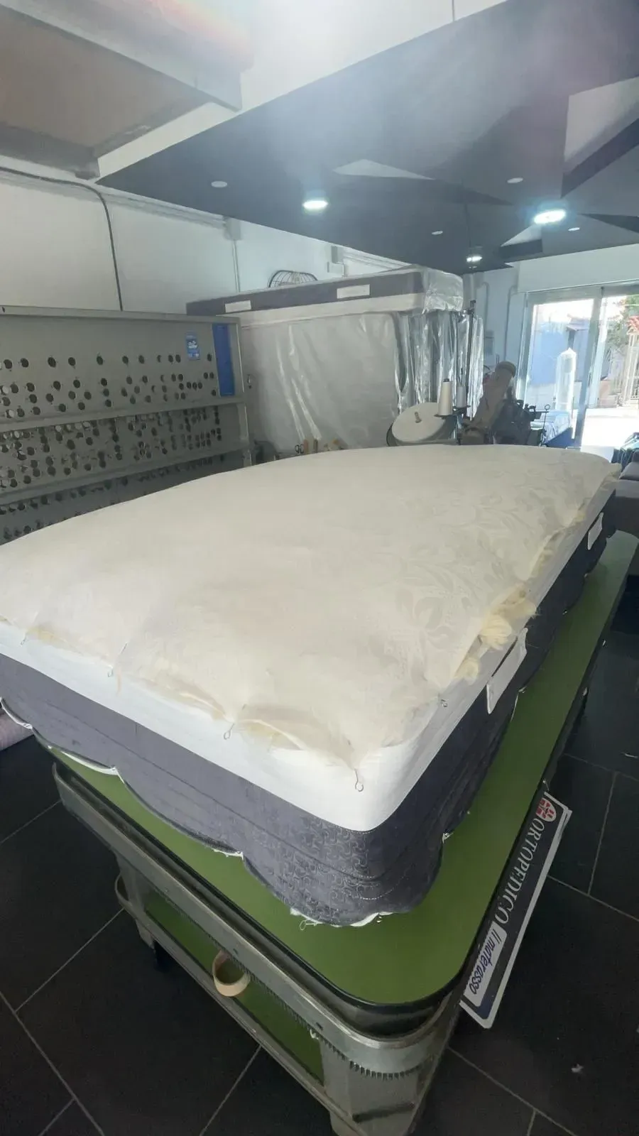 Un coprimaterasso bianco poggia su un materasso grigio in un laboratorio ben illuminato o in uno spazio commerciale con un soffitto nero.