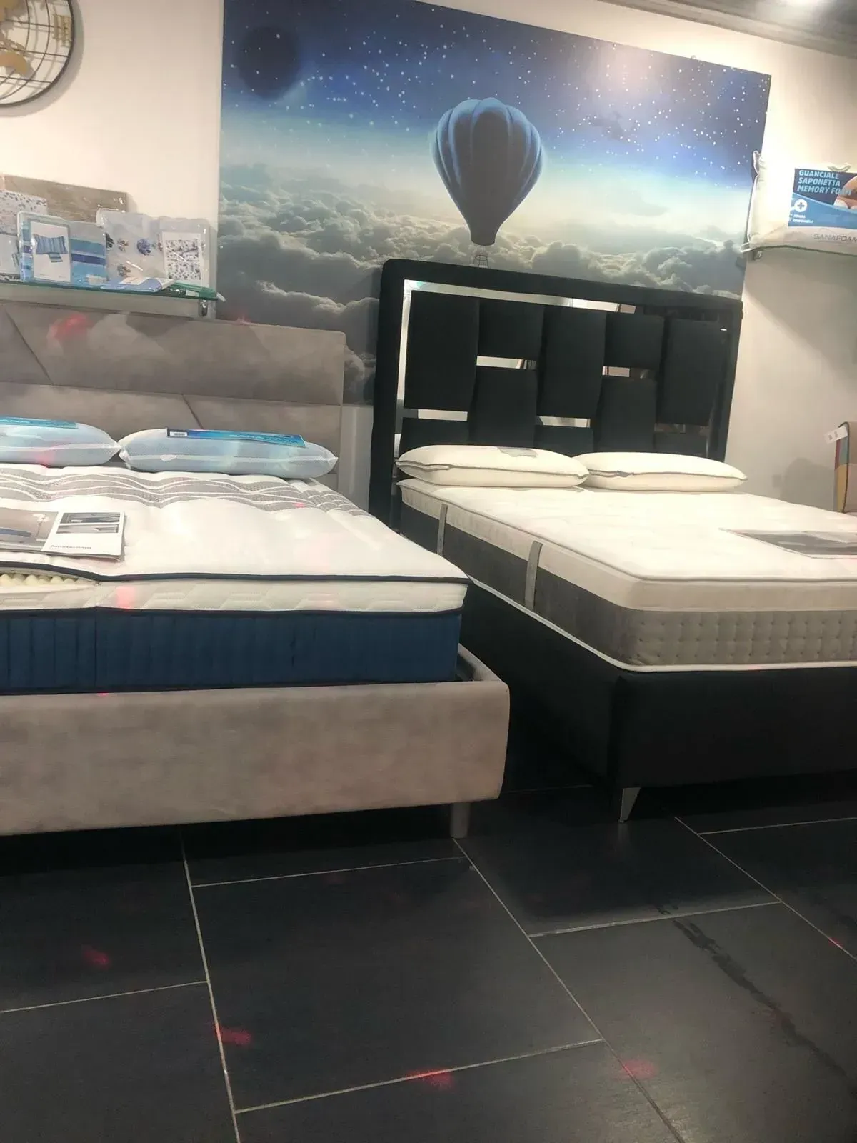 Due letti espositivi, uno grigio e uno nero, posizionati in uno showroom sotto un murale raffigurante una mongolfiera.