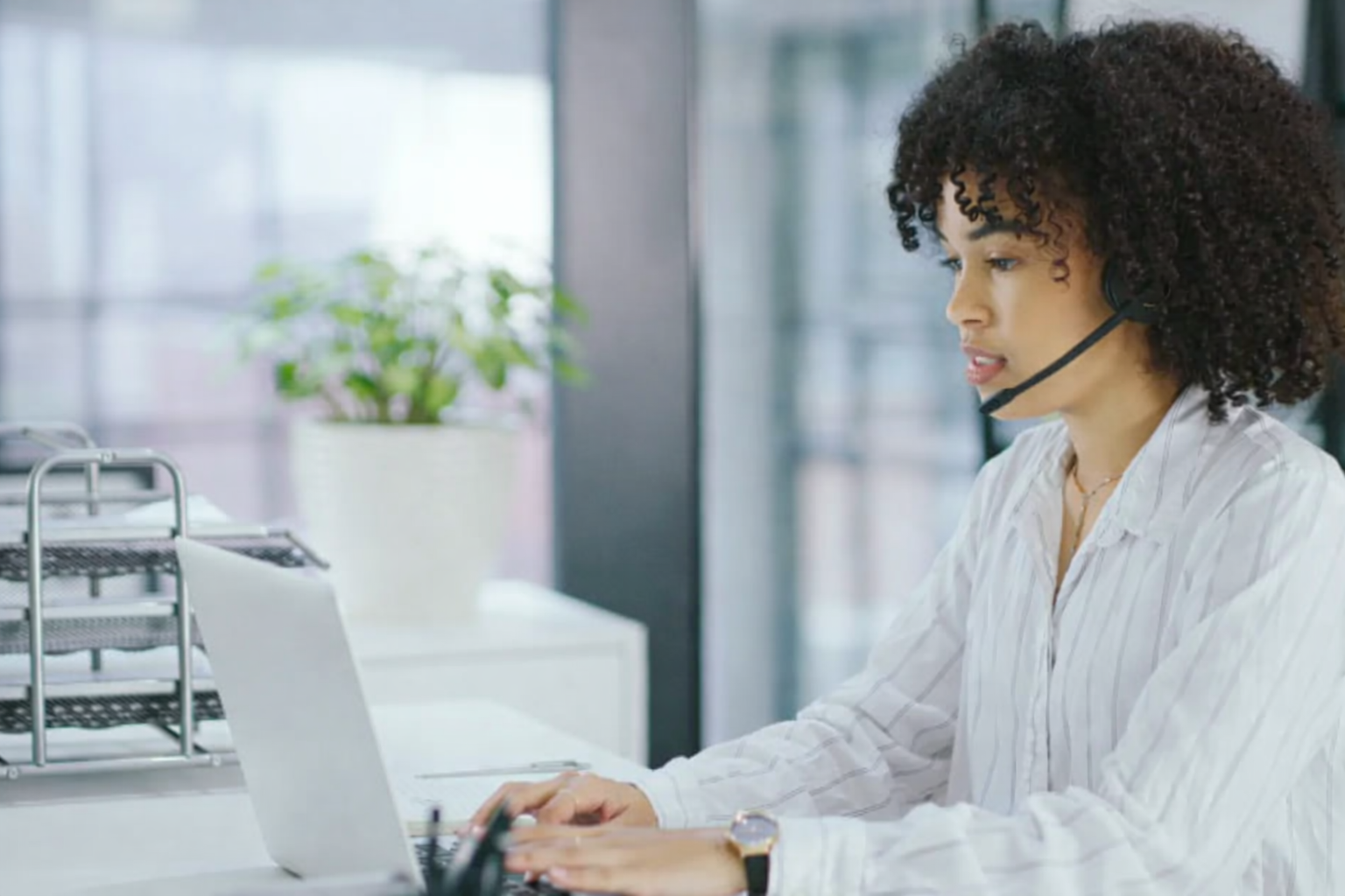 Woman wearing a headset, typing on a laptop at a desk in an office.