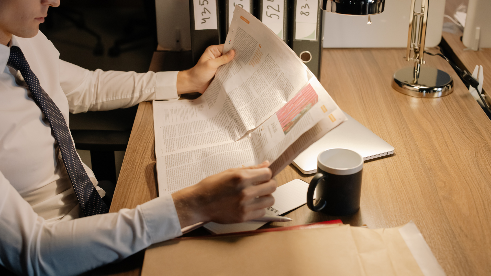 A man is sitting at a desk reading a newspaper.